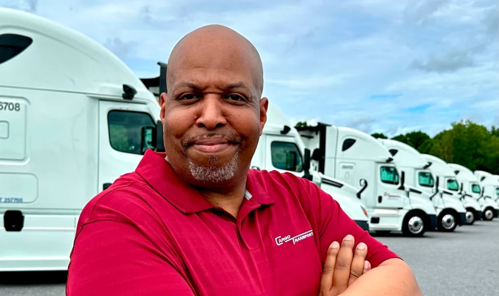 A professional CDL A truck driver in a red shirt standing confidently in front of white trucks, representing home weekly CDL jobs in Greater Charlotte NC.